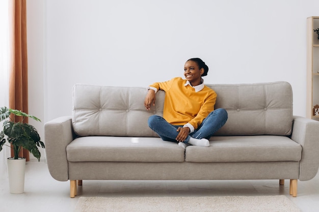Jovem negra aproveitando o fim de semana e relaxando no sofá confortável em casa Mulher afro-americana sentada no sofá na sala de estar esticando os braços e sorrindo, sentindo-se serena e feliz vista traseira
