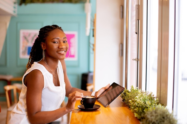 Jovem negra bebendo café enquanto olha para tablet digital no café