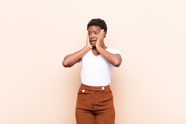 Jovem negra bonita se sentindo feliz, animada e surpresa, olhando para o lado com as duas mãos no rosto