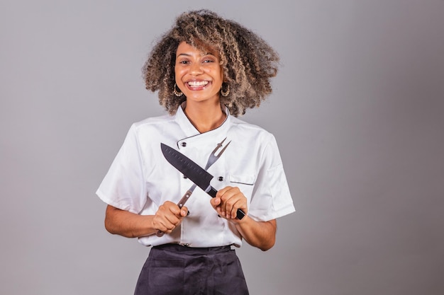 Foto jovem negra brasileira cozinheira masterchef vestindo uniforme de restaurante segurando garfo e faca para churrasco corte de carne