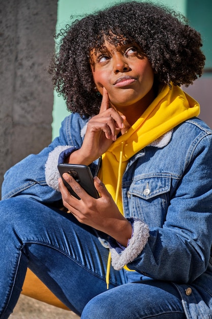 Jovem negra com cabelo afro sentada pensando com seu smartphone na mão