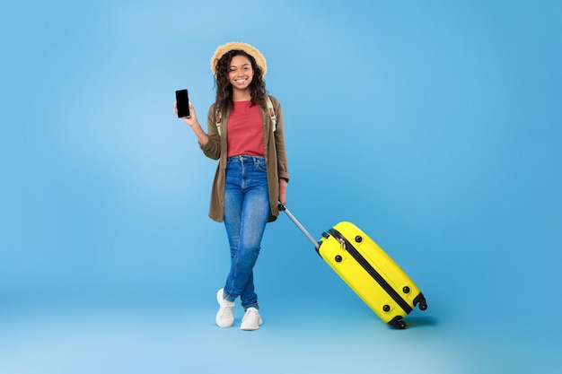 Jovem negra com mala mostrando smartphone com tela em branco oferecendo maquete reserva verão