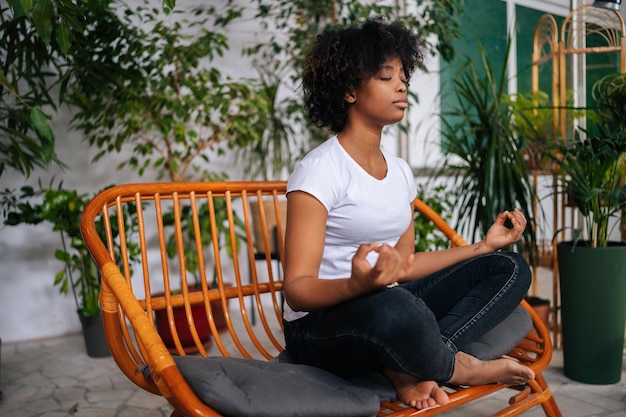 Jovem negra descalça consciente sentada com os olhos fechados no sofá em pose de lótus praticando ioga meditação em casa respirando fundo para a saúde mental