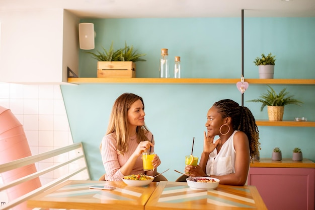 Jovem negra e caucasiana se divertindo bebendo sucos frescos e tomando café da manhã saudável no café