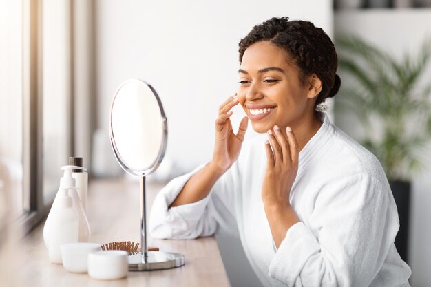 Foto jovem negra feliz aplicando hidratante facial enquanto olha no espelho de maquiagem