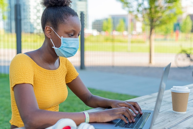 Jovem negra no trabalho em seu laptop em tempo de epidemia ao ar livre uso de máscara protetora jovem estudante no parque tomando seu café cappuccino a nova maneira normal de viver com proteção