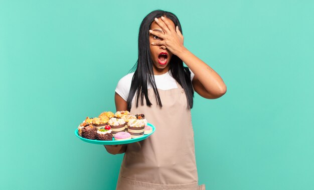 Jovem negra parecendo chocada, assustada ou apavorada, cobrindo o rosto com a mão e espiando por entre os dedos. conceito de chef de padaria