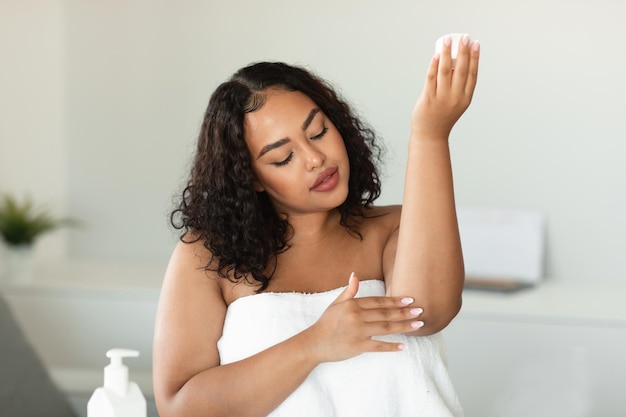 Jovem negra plus size em toalha aplicando creme calmante no cotovelo hidratando a pele seca em casa espaço livre