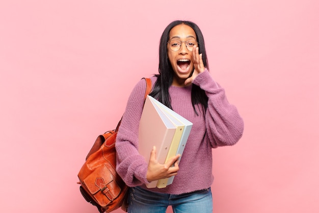 Jovem negra se sentindo feliz, animada e positiva, dando um grande grito com as mãos perto da boca, gritando. conceito de estudante