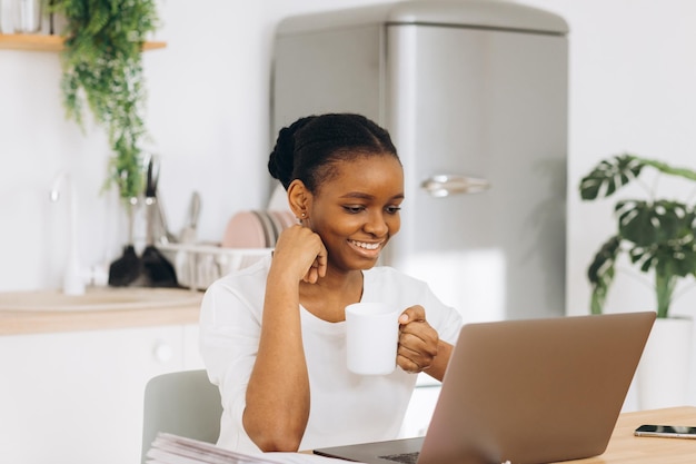 Jovem negra sentada na cozinha tomando café e falando em videochamada no laptop