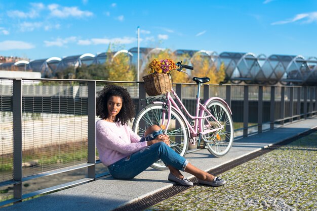 Foto jovem negra sentada perto do rio com sua bicicleta vintage