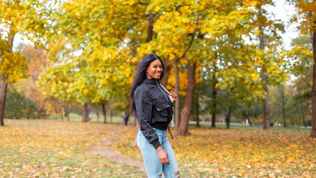 Jovem negra sorridente em roupas da moda casual com jaqueta elegante e jeans caminhando em um parque de outono com folhagem amarela de outono