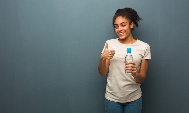 Jovem negra sorrindo e levantando o polegar, ela está segurando uma garrafa de água