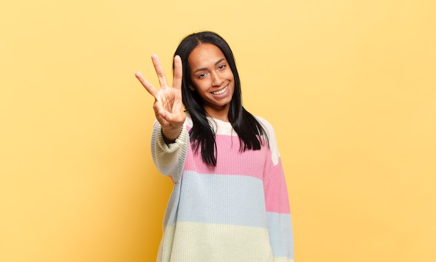 Jovem negra sorrindo e parecendo amigável, mostrando o número três ou o terceiro com a mão para a frente, em contagem regressiva