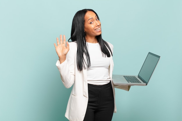 Jovem negra sorrindo feliz e alegre, acenando com a mão, dando as boas-vindas e cumprimentando ou dizendo adeus. conceito de laptop