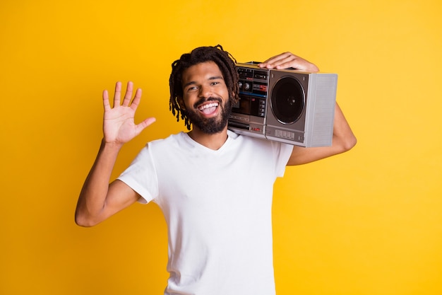 Jovem negro acenando com a mão segurando gravador vintage com a boca aberta e usar camiseta branca com fundo de cor amarela