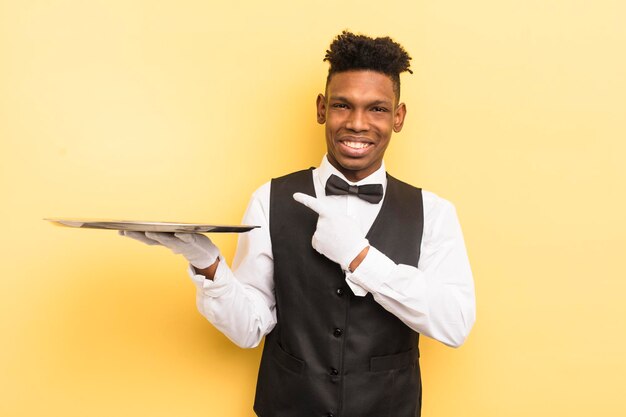 Jovem negro afro sorrindo alegremente se sentindo feliz e apontando para o conceito de garçom lateral com um