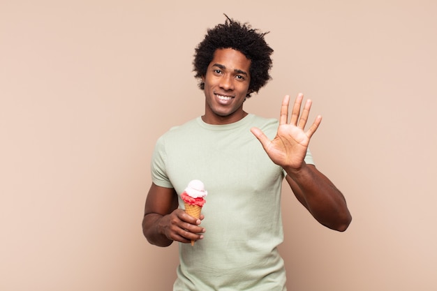 Jovem negro afro sorrindo e parecendo amigável, mostrando o número cinco ou quinto com a mão para a frente