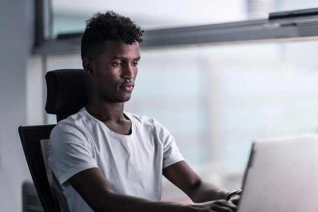 Jovem negro bonito concentrado focado em seu laptop e trabalhando com humor sério em um espaço de cópia de escritório moderno