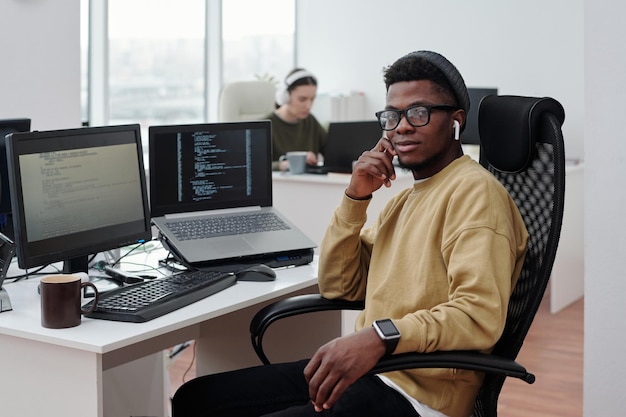 Jovem negro confiante em casualwear com fones de ouvido sentado no local de trabalho