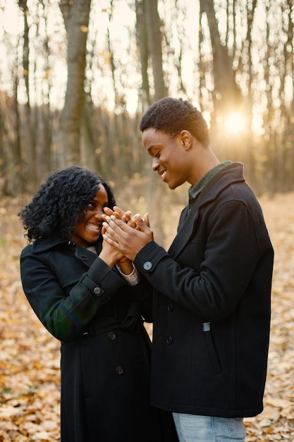 Jovem negro e sua namorada de mãos dadas. Casal romântico andando no parque outono no pôr do sol. Homem e mulher vestindo casacos pretos.