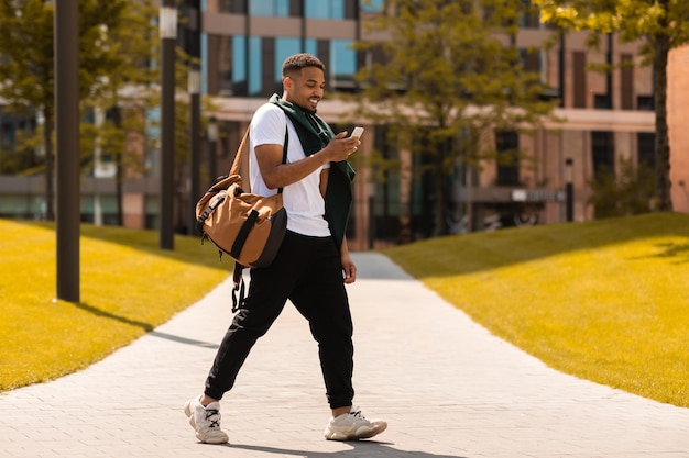 Jovem negro na moda andando na área urbana da cidade e mandando mensagens no smartphone ao ar livre