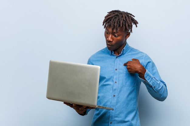 Jovem negro rasta segurando um laptop surpreendeu apontando para si mesmo, sorrindo amplamente.