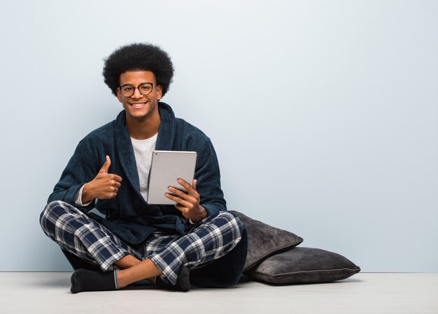 Jovem negro sentado em sua casa e segurando seu tablet sorrindo e levantando o polegar