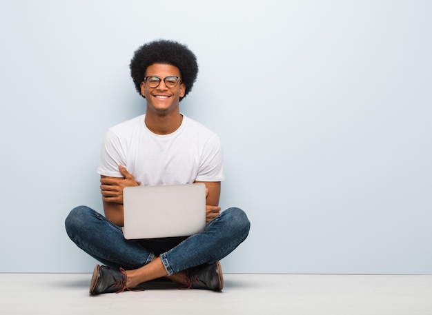 Jovem negro sentado no chão com um laptop cruzando os braços, sorrindo e relaxado