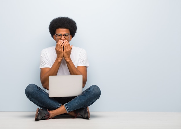 Jovem negro sentado no chão com um laptop muito assustado e com medo escondido