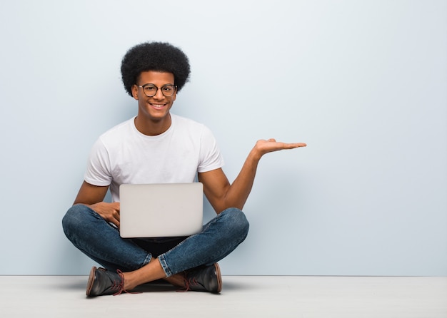Jovem negro sentado no chão com um laptop segurando algo com a mão