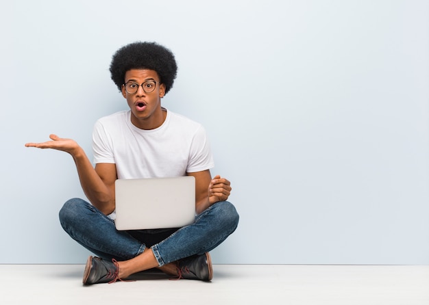 Jovem negro sentado no chão com um laptop segurando algo na palma da mão