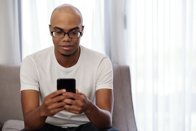 Jovem negro sorridente de óculos, sentado no sofá e lendo as notícias nas redes sociais
