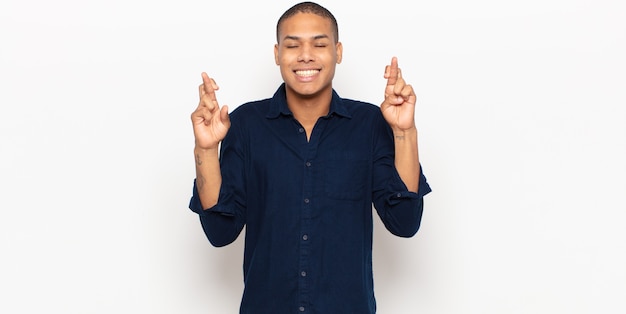 Jovem negro sorrindo e cruzando os dedos ansiosamente, sentindo-se preocupado e desejando ou esperando por boa sorte