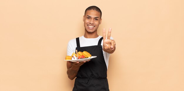 Jovem negro sorrindo e parecendo feliz, despreocupado e positivo, gesticulando vitória ou paz com uma mão