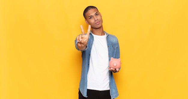 Jovem negro sorrindo e parecendo feliz, despreocupado e positivo, gesticulando vitória ou paz com uma mão