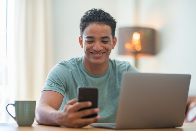 Jovem negro usando conexão de telefone e computador em casa homem feliz lendo notificação no celular móvel enquanto trabalha no laptop em trabalho inteligente pessoas modernas da geração z usando web