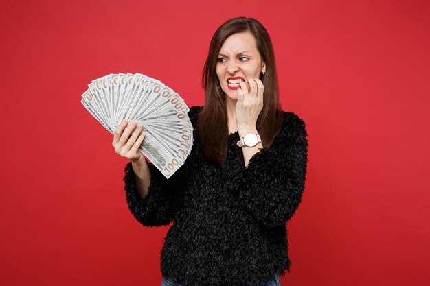 Jovem nervosa de suéter de pele preta roendo as unhas, segurando um leque de dinheiro nas notas de dólar, dinheiro isolado sobre fundo vermelho. Emoções sinceras de pessoas, conceito de estilo de vida. Simule o espaço da cópia.