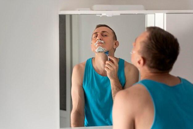 Jovem no banheiro remove a barba com espuma de barbear no rosto Cuidados com o rosto Reflexão no espelho