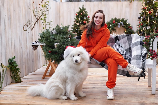 Jovem no fundo da árvore de Natal com cachorro samoiedo branco no chapéu de Papai Noel ao ar livre Decoração de quintal para o Ano Novo