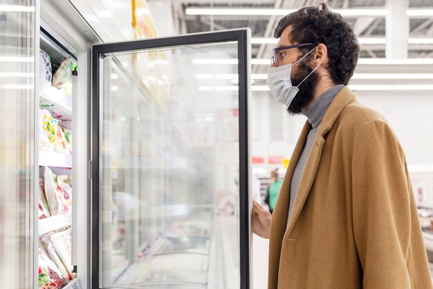 Jovem no supermercado no departamento com alimentos congelados. uma morena com uma máscara médica durante a pandemia de coronavírus.