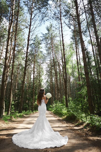 Jovem noiva elegante com joias no penteado em vestido de noiva de renda, mantendo um buquê e posando na floresta.