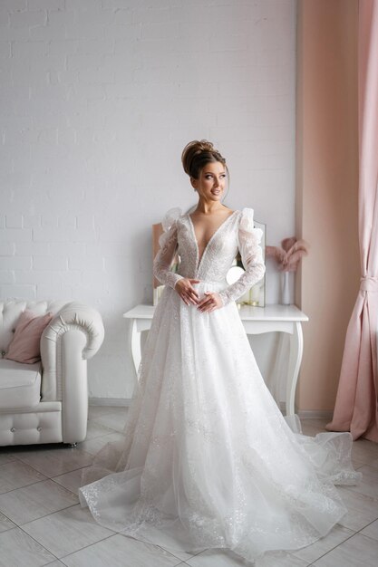 Jovem noiva em um vestido de noiva branco posando em um interior claro foto de corpo inteiro cabelo castanho maquiagem profissional e foto de penteado para um salão de casamento ou agência