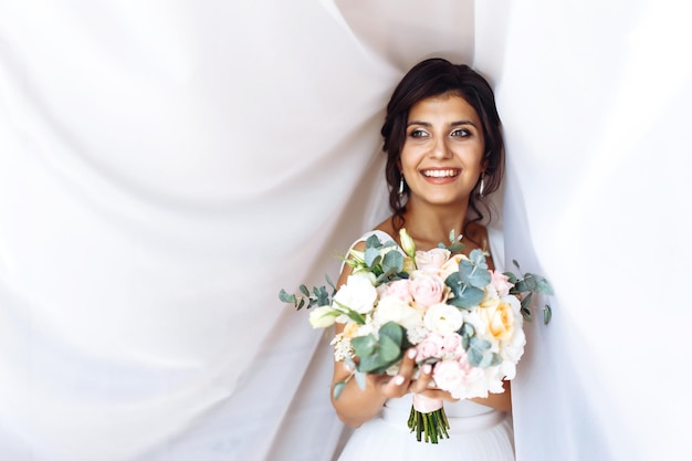 Foto jovem noiva em vestido branco com um buquê de flores posando sob a cortina manhã da noiva