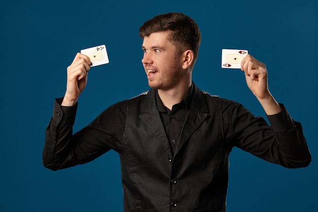 Jovem, novato no pôquer, de colete e camisa pretos. segurando duas cartas de jogar e parecendo animado. posando contra o fundo azul do estúdio. jogos de azar, cassino. fechar-se.