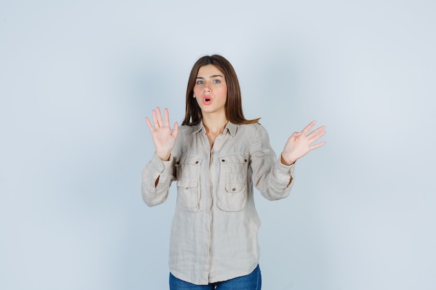 Jovem numa camisa bege, jeans levantando as mãos como mostrando gesto de restrição e parecendo surpresa, vista frontal.