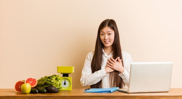 Jovem nutricionista chinesa trabalhando com seu laptop tem uma expressão amigável, pressionando a palma da mão no peito. Conceito de amor.