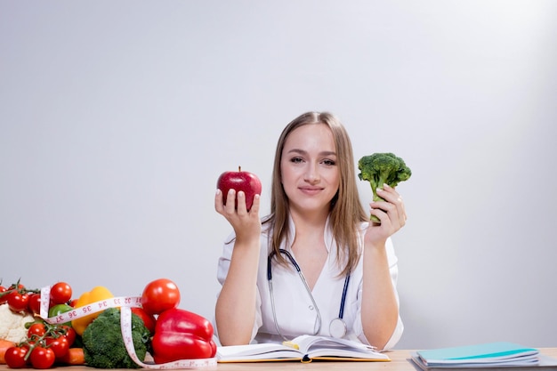 Jovem nutricionista positiva sentada à mesa e segurando brócolis e uma maçã nas mãos Conceito de alimentação saudável