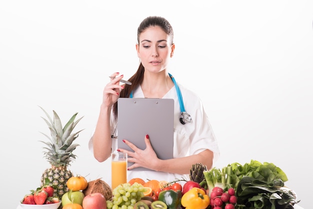 Jovem nutricionista sentada à mesa e mostrando frutas e vegetais coloridos