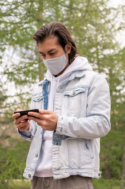 Jovem ocupado em roupas casuais e máscara protetora assistindo vídeo no smartphone